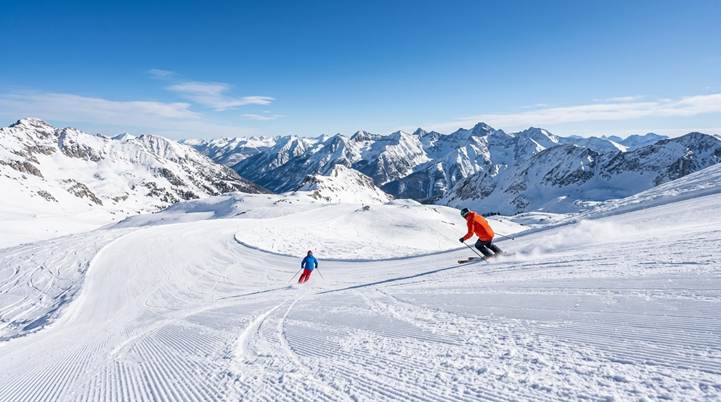 Two skiers enjoy a sunny day on a wide, groomed run at a high-altitude Colorado ski area, with the Rocky Mountains in the background.